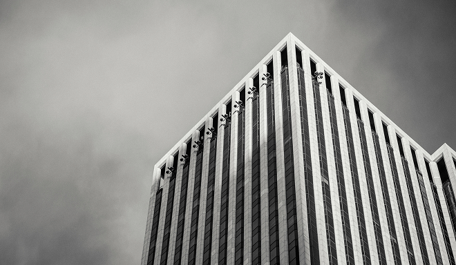 Fotografía en blanco y negro de un edificio de oficinas moderno de gran altura visto desde la calle, con líneas verticales y cielo nublado